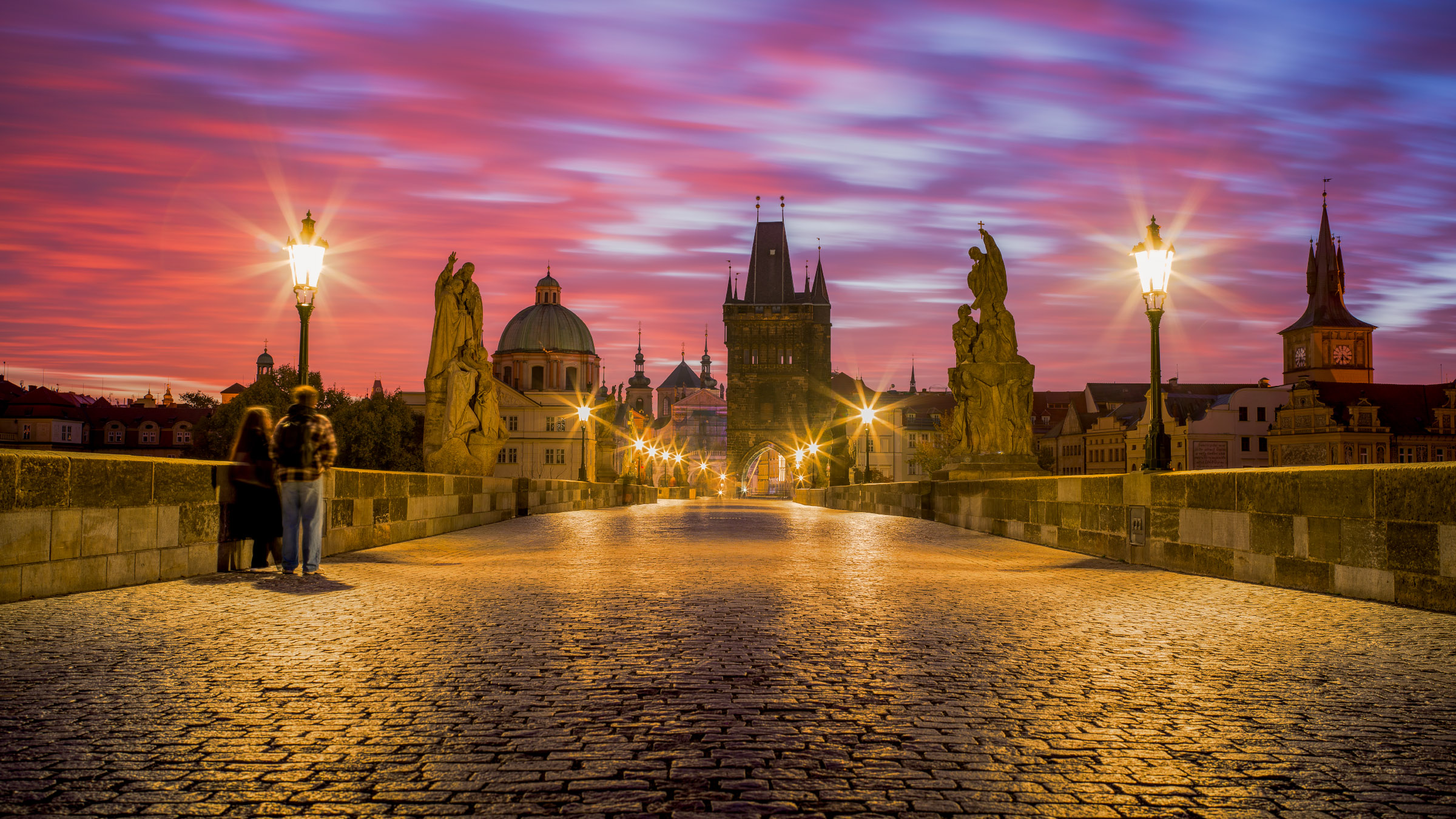 Cobbled Charles Bridge with glowing lamps, statues, and two blurred figures under a streaked pink dawn sky, Prague.