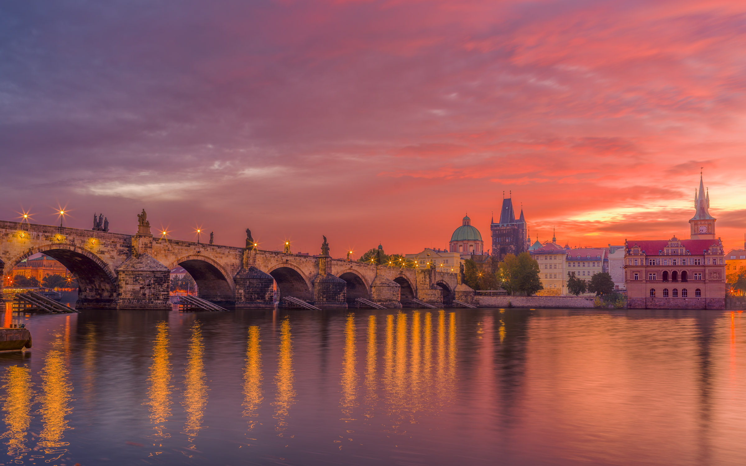 Charles Bridge with lit lamps reflected in the Vltava River under a pink and orange dawn sky, Old Town towers at right, Prague.