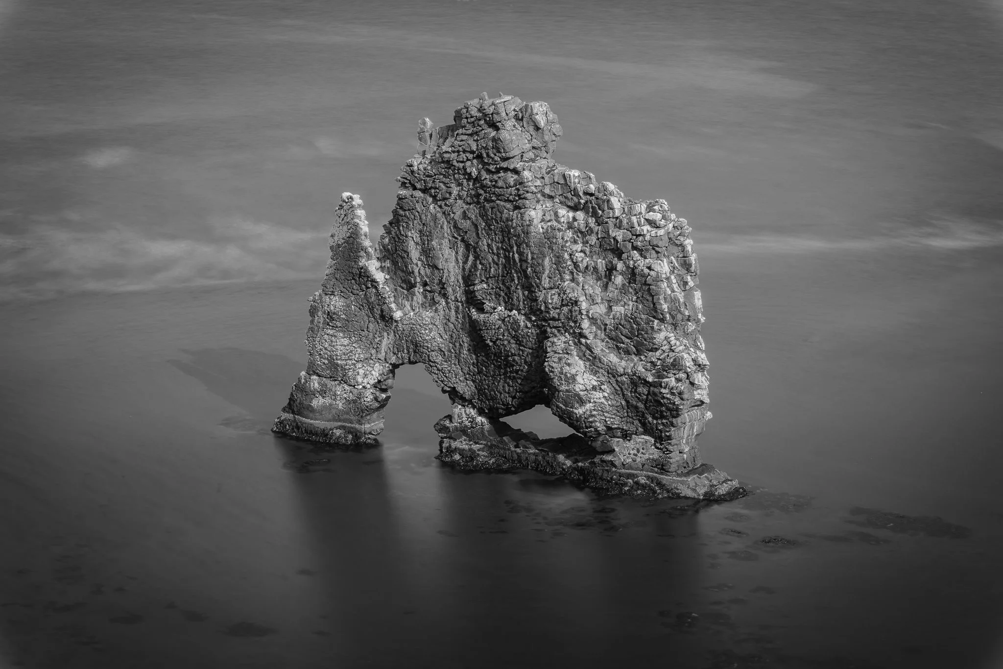 Hvítserkur rock formation in Iceland captured in black and white long exposure, creating a dramatic silhouette against calm waters and cloudy sky.