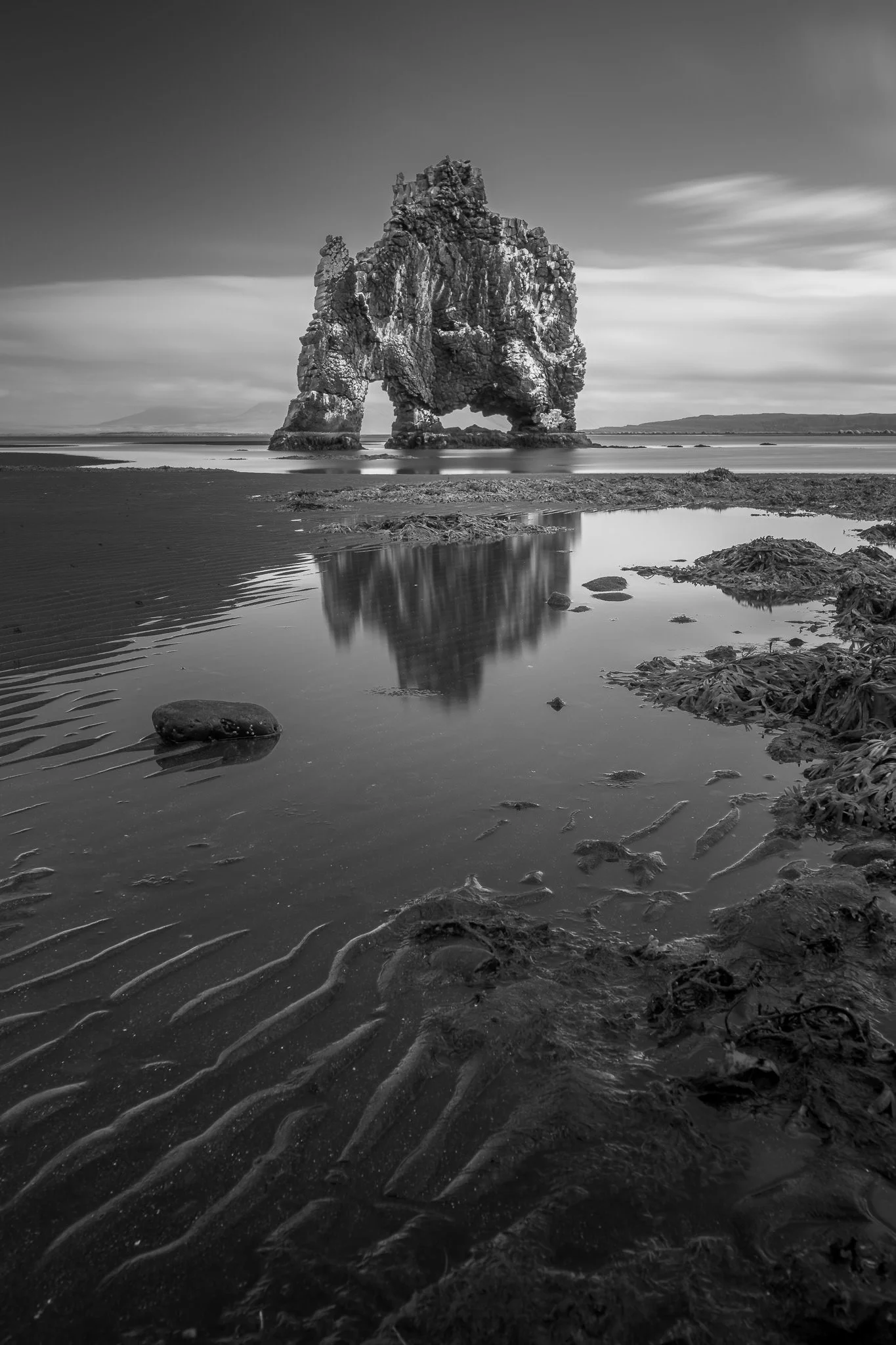 Hvítserkur rock formation in Iceland captured in black and white long exposure, creating a dramatic silhouette against calm waters and cloudy sky.